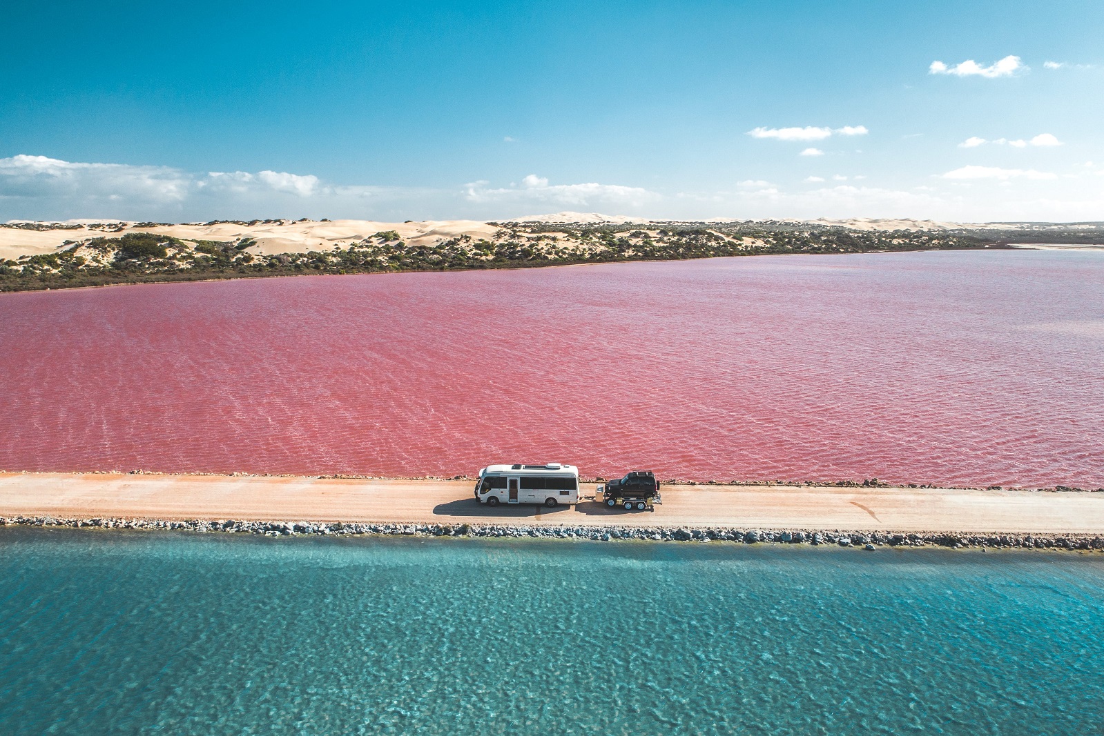 A white tour bus drives past the Pink Lake on South Australia's Eyre Peninsula. It is a bright, sunny day with the dazzling pink waters of the lake, sparkling in the sunshine.
